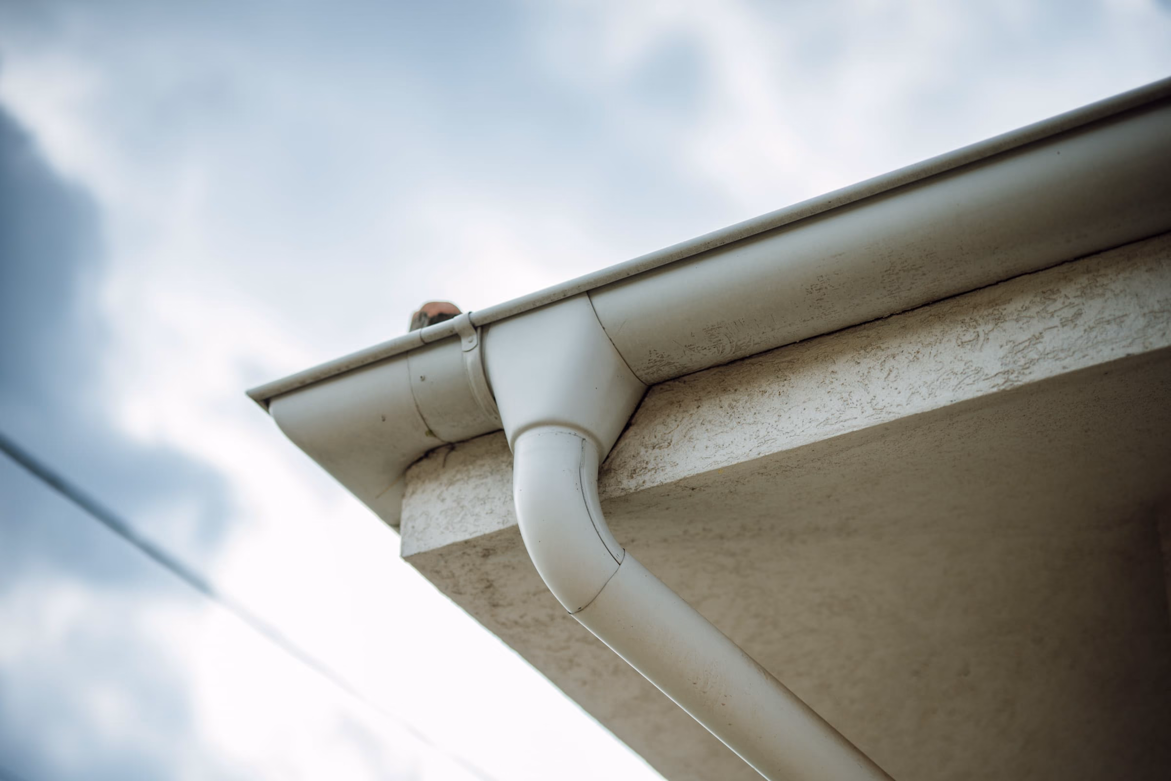 A white K-style gutter meeting a downspout elbow at the corner of a roof eave, photographed from below against an overcast sky.
