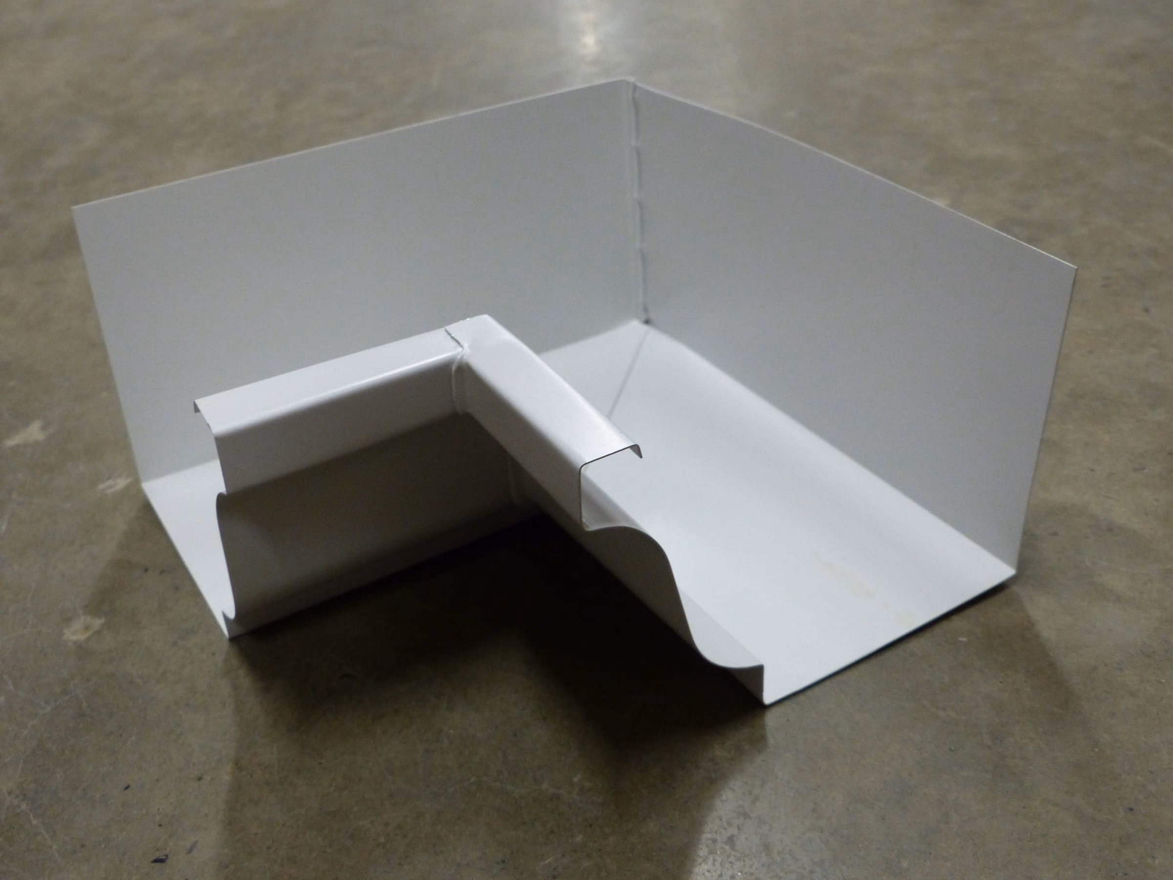 A white K-style gutter corner mitre resting on a concrete floor, showing the ogee profile of the gutter front.
